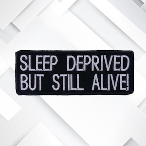 Peut inclure: Écusson brodé noir avec du texte blanc qui dit "SLEEP DEPRIVED BUT STILL ALIVE!"