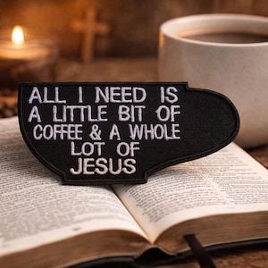 May include: A black patch in the shape of a coffee cup with white embroidered text reading "ALL I NEED IS A LITTLE BIT OF COFFEE & A WHOLE LOT OF JESUS" sits on an open Bible. A white mug of coffee and a lit candle are in the background.