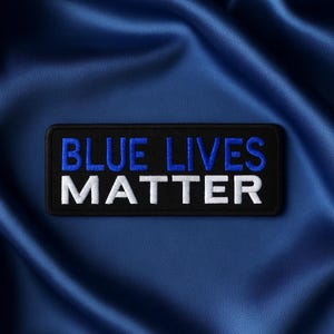 Puede incluir: Un parche rectangular negro con el texto bordado "BLUE LIVES MATTER" en azul y blanco. El parche está sobre un fondo de tela azul.