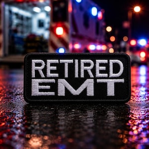 May include: A black patch with white embroidered text that reads "RETIRED EMT." The patch is rectangular and sits on a wet, reflective surface. An ambulance with flashing lights is blurred in the background.