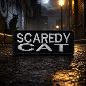 Pode incluir: Um patch retangular preto com as palavras "SCAREDY CAT" bordadas em branco. O patch está situado em uma cena de rua escura e molhada com a silhueta de um gato e uma lua cheia ao fundo.