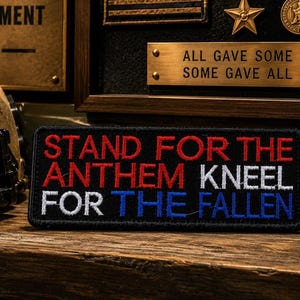 May include: A black patch with red, white, and blue embroidered text that reads "STAND FOR THE ANTHEM KNEEL FOR THE FALLEN." The patch is on a wooden surface, with a military helmet and other military memorabilia in the background.