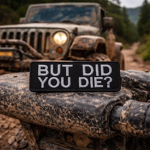 Puede incluir: Un parche rectangular negro con texto bordado blanco que dice "BUT DID YOU DIE?". El parche está colocado sobre un vehículo embarrado, con un Jeep al fondo. La imagen está tomada al aire libre.
