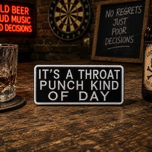 May include: A black patch with white text saying "IT'S A THROAT PUNCH KIND OF DAY." The patch is on a wooden surface. A glass of amber liquid, a bottle with a skull label, and a neon sign are in the background.