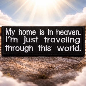 May include: A black rectangular patch with white embroidered text that reads, "My home is in heaven. I'm just traveling through this world." A silver cross pendant hangs in the background.