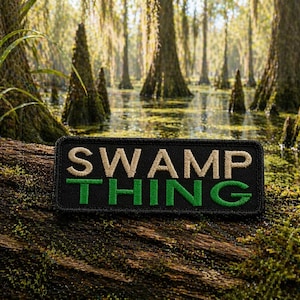May include: A black rectangular patch with the words "SWAMP THING" embroidered in tan and green. The patch is set against a swampy background with trees and water.