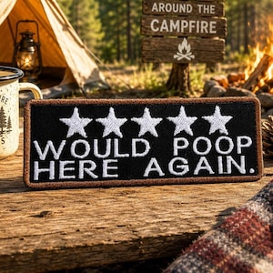 May include: A black patch with white embroidered text and five white stars. The text reads "WOULD POOP HERE AGAIN." The patch is on a wooden surface, with a campfire and tent in the background. A mug with "ADVENTURE AWAITS" is also visible.