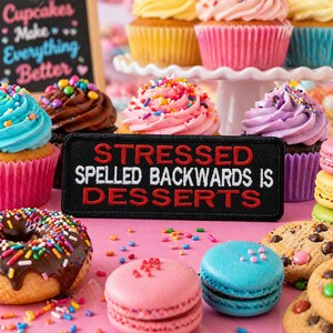 May include: A colorful assortment of cupcakes, donuts, and macarons on a pink surface. The cupcakes have various frosting colors and sprinkles. A black patch reads "STRESSED SPELLED BACKWARDS IS DESSERTS."