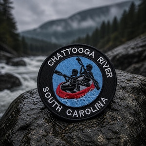May include: A round patch with the text "Chattooga River South Carolina" encircling a blue center. The center features a red canoe with two figures paddling. The patch is black with white lettering, set against a blurred river and mountain backdrop.