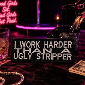 Peut inclure: Un patch noir avec du texte blanc brodé qui dit "I WORK HARDER THAN A UGLY STRIPPER." L'image comprend également un verre de whisky, des talons hauts et une enseigne au néon qui dit "Good Girls Sit. Bad Girls Get Paid."