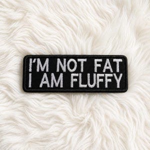 Könnte beinhalten: Schwarzer rechteckiger Aufnäher mit weiß gesticktem Text: „I'M NOT FAT I AM FLUFFY“. Der Aufnäher befindet sich vor einem weißen, flauschigen Hintergrund.