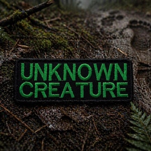 May include: A black rectangular patch with the words "UNKNOWN CREATURE" embroidered in bright green. The patch is set against a backdrop of forest floor with leaves and ferns.
