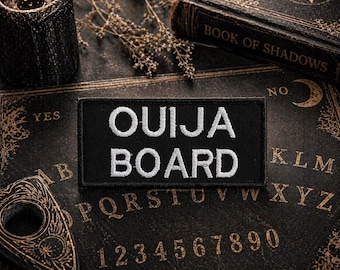 May include: A black Ouija Board patch with white embroidered text reading "OUIJA BOARD" rests on a vintage Ouija board. A lit black candle, a planchette, and a book titled "BOOK OF SHADOWS" are also visible, creating a mystical theme.