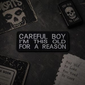 May include: A black patch with white embroidered text that reads "CAREFUL BOY I'M THIS OLD FOR A REASON." The image also includes a skull lighter, a notebook, and a Misfits patch.