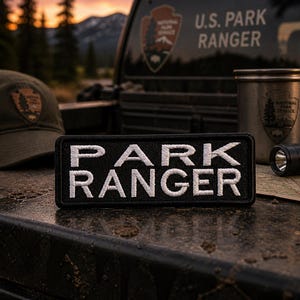 May include: A black rectangular patch with white embroidered text that reads "PARK RANGER." The patch is displayed on a textured surface, with a hat, a metal cup, and a flashlight in the background. The image has a National Park Service theme.