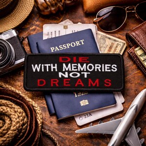 Può includere: Una toppa nera con la scritta "DIE WITH MEMORIES NOT DREAMS" in bianco e rosso, appoggiata su passaporti blu. L'immagine include una macchina fotografica, occhiali da sole, un portafoglio e un aereo giocattolo, suggerendo un tema di viaggio.