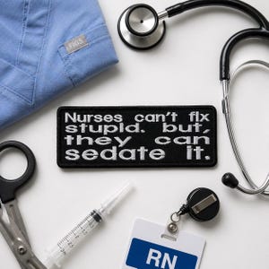 Puede incluir: Un parche negro bordado con texto blanco que dice "Nurses can't fix stupid, but they can sedate it." También son visibles herramientas médicas, incluyendo un estetoscopio, una jeringa, tijeras y una etiqueta de identificación que dice "RN."