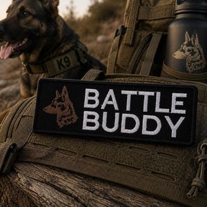 May include: A black patch with the words "BATTLE BUDDY" in white, and a brown dog silhouette. A black water bottle with a dog silhouette is in the background. A German Shepherd dog is also in the background, wearing a "K9" collar.
