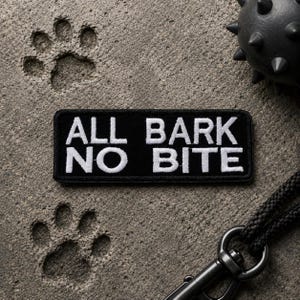 May include: A black rectangular patch with the text "ALL BARK NO BITE" in white lettering. The patch is on a textured gray surface with paw prints and a black dog toy.