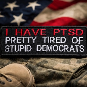 Op de afbeelding: Een zwart rechthoekig embleem met witte tekst die luidt: "I HAVE PTSD PRETTY TIRED OF STUPID DEMOCRATS" tegen een camouflage achtergrond met een Amerikaanse vlag op de achtergrond.