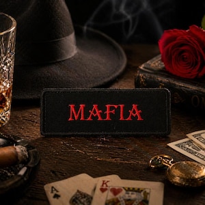 Puede incluir: Naturaleza muerta con un sombrero fedora negro, un vaso de líquido ámbar, un cigarro encendido en un cenicero, naipes, un reloj de bolsillo y una rosa roja. También hay un parche negro con la palabra "MAFIA" en letras rojas.