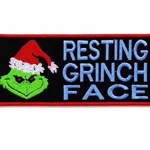 May include: Embroidered patch with a black background and a red border. The patch features a green Grinch wearing a Santa hat and the words "RESTING GRINCH FACE" in blue.
