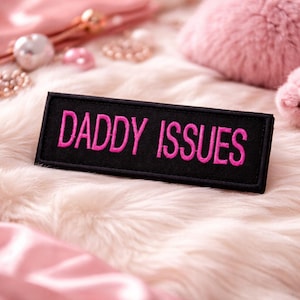 May include: A black rectangular patch with the embroidered text "DADDY ISSUES" in bright pink. The patch is set against a backdrop of fluffy, light-colored faux fur, with pink accessories in the background.