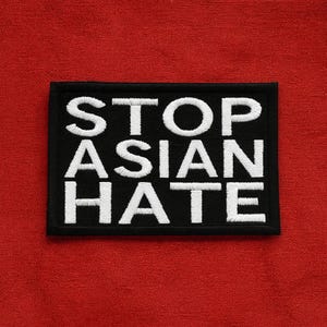 May include: A rectangular black patch with white embroidered text that reads "STOP ASIAN HATE." The patch is set against a red background. The text is in a bold, sans-serif font.
