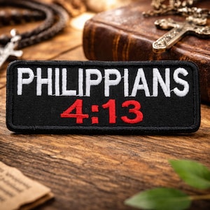 May include: A black patch with the text "PHILIPPIANS 4:13" in white and red. The patch is on a wooden surface, with a rosary, a leather-bound book, and a cross necklace in the background. Green leaves and a piece of paper are also visible.