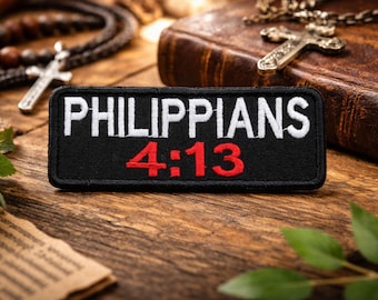 以下が含まれることがあります： 白と赤で「PHILIPPIANS 4:13」と書かれた黒いパッチ。木製の表面にあり、ロザリオ、革張りの本、十字架のネックレスが背景にあります。緑の葉と紙も表示されています。