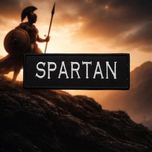 Puede incluir: Parche rectangular negro con la palabra "SPARTAN" bordada en blanco. La imagen muestra un guerrero espartano con lanza y escudo, con un fondo de puesta de sol sobre un paisaje montañoso.
