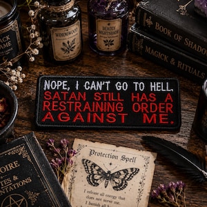 Może przedstawiać: Czarna, haftowana naszywka z napisem "NOPE, I CAN'T GO TO HELL. SATAN STILL HAS A RESTRAINING ORDER AGAINST ME." jest otoczona świecami, książkami i butelkami z miksturami. Scena ma gotycki lub czarowniczy motyw.