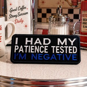 May include: A black embroidered patch with white text that reads "I HAD MY PATIENCE TESTED I'M NEGATIVE." The patch is on a diner table next to a coffee mug and condiments.