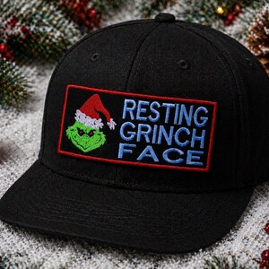 May include: Black baseball cap with a rectangular patch featuring the Grinch wearing a Santa hat and the words "RESTING GRINCH FACE" in blue lettering. The patch is outlined in red. The cap is surrounded by Christmas decorations.