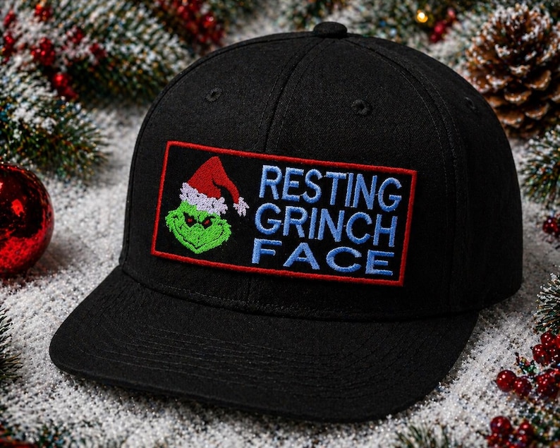 May include: Black baseball cap with a rectangular patch featuring the Grinch wearing a Santa hat and the words "RESTING GRINCH FACE" in blue lettering. The patch is outlined in red. The cap is surrounded by Christmas decorations.