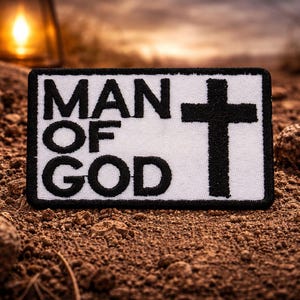 May include: A rectangular patch with the words "MAN OF GOD" in bold black letters and a black cross on a white background. The patch is set on a bed of brown soil with a blurred background.