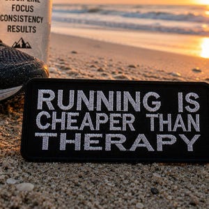 Peut inclure: Écusson noir avec texte brodé blanc « RUNNING IS CHEAPER THAN THERAPY » sur une plage de sable. Chaussures de course noires et un récipient blanc avec du texte en arrière-plan. Le ciel est de couleur orange chaud.