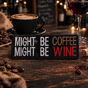 May include: A black patch with white, brown, and red embroidered text that reads "MIGHT BE COFFEE MIGHT BE WINE." The patch is on a wooden surface, with a cup of coffee, a glass of red wine, and coffee beans in the background.