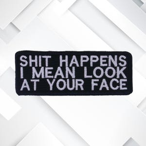 May include: Black rectangular patch with white embroidered text that reads "SHIT HAPPENS I MEAN LOOK AT YOUR FACE". The patch is likely designed to be sewn or ironed onto clothing or accessories.