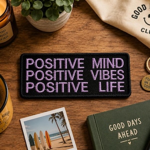 May include: A black embroidered patch with the words "POSITIVE MIND POSITIVE VIBES POSITIVE LIFE" in purple. The image also includes a coffee mug, a candle, a small plant, a keychain, a notebook, and a photo of surfboards on a beach.