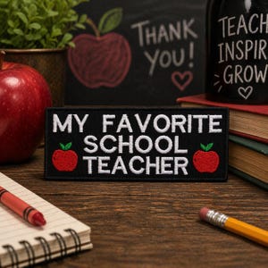 Könnte beinhalten: Ein schwarzer Aufnäher mit den Worten "MY FAVORITE SCHOOL TEACHER" in Weiß. Rote Apfelgrafiken befinden sich auf beiden Seiten des Textes. Ein roter Buntstift, ein Bleistift und ein Notizblock befinden sich im Vordergrund. Eine Tafel mit "Thank You!" befindet sich im Hintergrund.