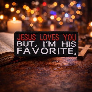 Könnte beinhalten: Ein schwarzer rechteckiger Aufnäher mit dem Text "JESUS LOVES YOU BUT, I'M HIS FAVORITE" in roter und weißer Stickerei. Der Aufnäher wird auf einer Holzoberfläche mit einem verschwommenen Hintergrund aus Lichtern und einer Kerze angezeigt.