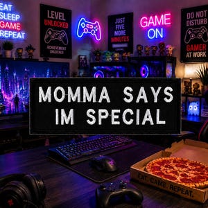 May include: A gamer's room with neon signs that read "Eat Sleep Game Repeat", "Level Unlocked", and "Game On". A black patch says "Momma Says I'm Special". A pizza and gaming accessories are on the desk.