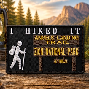 May include: Black rectangular patch with white text and a hiker graphic. Text reads "I Hiked It Angels Landing Trail Zion National Park 4.4 Miles". The patch is on a wooden surface with a mountain backdrop.