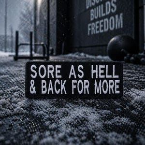 Op de afbeelding: Een zwart rechthoekig embleem met witte geborduurde tekst "SORE AS HELL & BACK FOR MORE". Het embleem ligt op een getextureerd oppervlak met een besneeuwde achtergrond. De achtergrond bevat trainingsapparatuur en de woorden "DISCIPLINE BUILDS FREEDOM".
