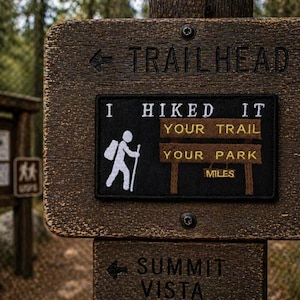 May include: A rustic wooden signpost with "TRAILHEAD" and an arrow carved into it. A black patch displays "I HIKED IT YOUR TRAIL YOUR PARK MILES" and a hiker icon. Below, another sign points to "SUMMIT VISTA".
