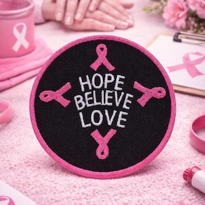 May include: A round black patch with a pink border and the words "HOPE BELIEVE LOVE" embroidered in white. Pink ribbons are embroidered around the words. The patch is on a pink surface, with other pink items in the background.