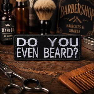 May include: A black embroidered patch with the text "DO YOU EVEN BEARD?" in white. The patch is displayed on a wooden surface with barber tools and beard care products in the background.