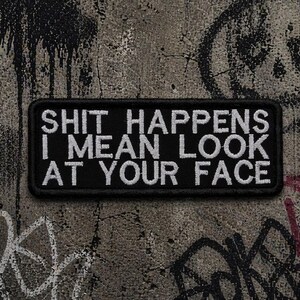 May include: A black rectangular patch with white embroidered text that reads "SHIT HAPPENS I MEAN LOOK AT YOUR FACE." The patch is set against a textured, graffiti-covered concrete wall.
