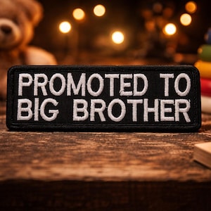 May include: A black patch embroidered with the words "PROMOTED TO BIG BROTHER" in white. The patch is surrounded by children's toys, including wooden blocks, a teddy bear, and a stacking ring toy.